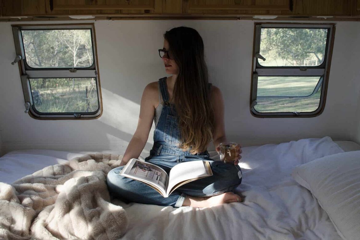 Frau mit einem Tee und Buch im Bett eines Tiny House, in dem sie übernachtet